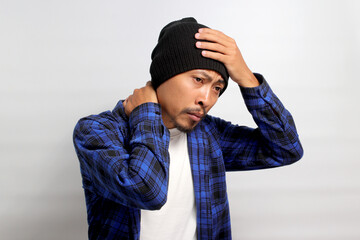 Young Asian man, dressed in a beanie hat and casual shirt, is holding his head, suggesting he's experiencing a headache, isolated on a white background. Healthcare and medical concepts