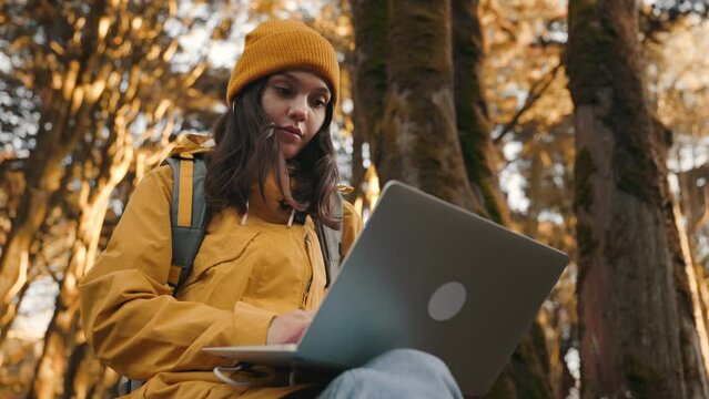 A young woman in a yellow coat and hat enjoys remote work in a serene forest, typing on a laptop in a peaceful natural setting.