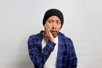 Young Asian man in a beanie hat and casual shirt is is suffering from a toothache, experiencing discomfort or pain in his tooth, isolated on white background. Healthcare and medical concepts