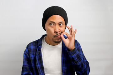Young Asian man, dressed in a beanie hat and casual shirt, zips his lips in a promise to keep a...