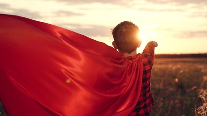 Boy shows superhero character pose in field wearing vibrant cloak. Boy with cloak fluttering...