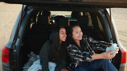 Happy multicultural female friends take selfie on camera in trunk of car by estuary on sand in summertime