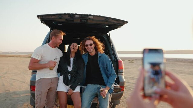 Sunny multinational friends taking cell phone pictures with beer in trunk of car near estuary