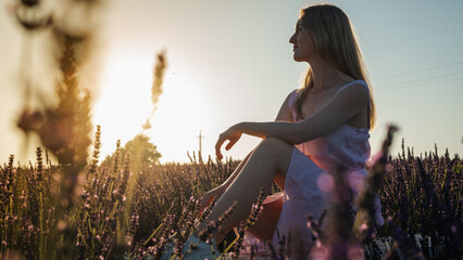 counter portrait of a young girl in a white summer dress on a lavender field, looks from behind, raised her hands to the top of the sun. Blooming lavender in summer. Sunset. Selective focus