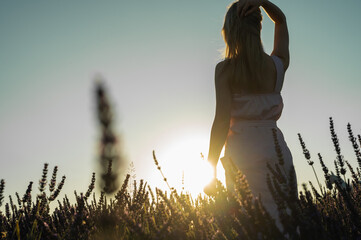 counter portrait of a young girl in a white summer dress on a lavender field, looks from behind, raised her hands to the top of the sun. Blooming lavender in summer. Sunset.