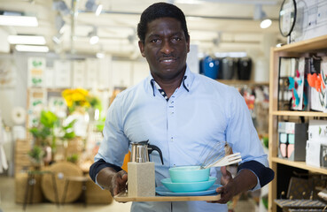 Satisfied african american male shopper holding dishware and kitchen accessories bought in...