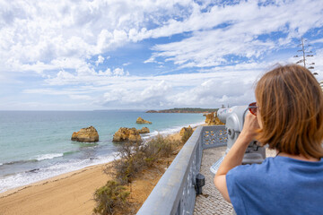 Woman looking at the sea from a binocular to the coast of Algarve. Beach of the Tree Castles (Praia...