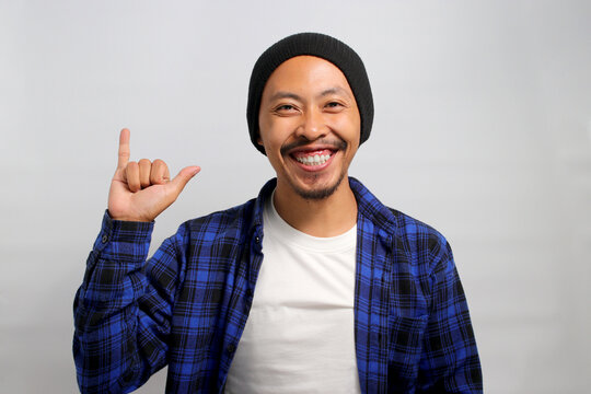 Young Asian man, wearing a beanie hat and casual clothes, flashes a rock gesture and a smile at the camera, showing his excitement and readiness to attend his favorite bands concert, white background