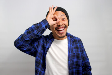 Excited young Asian man, wearing a beanie hat and casual shirt, is making an OK gesture with his hand positioned as if using binoculars, with his eyes peering through the opening formed by his fingers