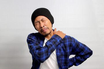Asian man, dressed in a beanie hat and casual outfit, is suffering from neck and shoulder pain,...