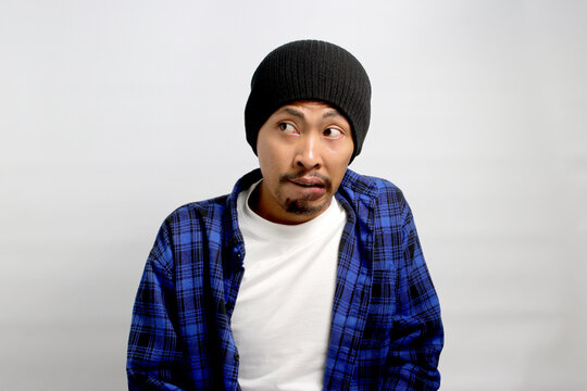 Hesitant young Asian man, dressed in a beanie hat and casual shirt, bites his lips, conveying a playful oops moment as he looks away towards empty space, against a white background
