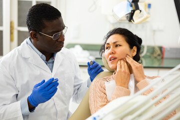 Asian woman patient talking to African American male dentist and complaining about toothache at dental clinic office.. - obrazy, fototapety, plakaty