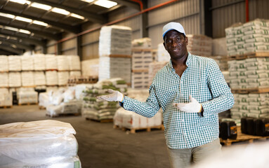 Portrait of positive African American man worker posing at warehouse, pointing something