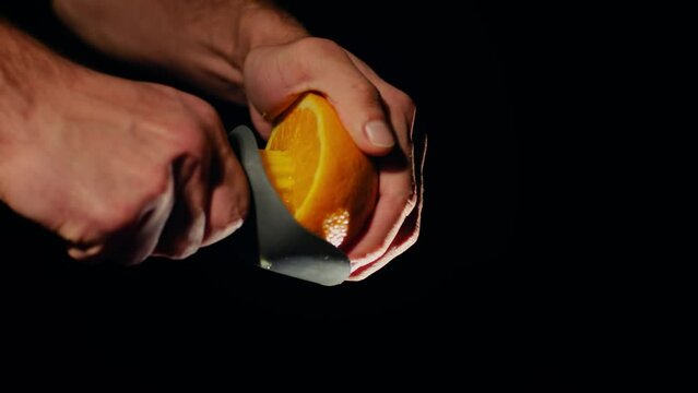 Bartender is squeezing juice from an orange. Closeup of chef’s hand squeezing juice from an orange by a hand squeezer on black background. Slowmotion, footage 4K. 