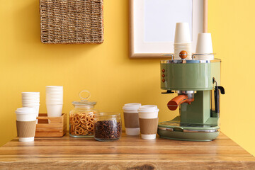 Modern coffee machine, takeaway cups and snacks on table near yellow wall