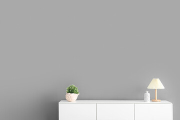 Wooden chest of drawers with houseplant and lamp near grey wall in room