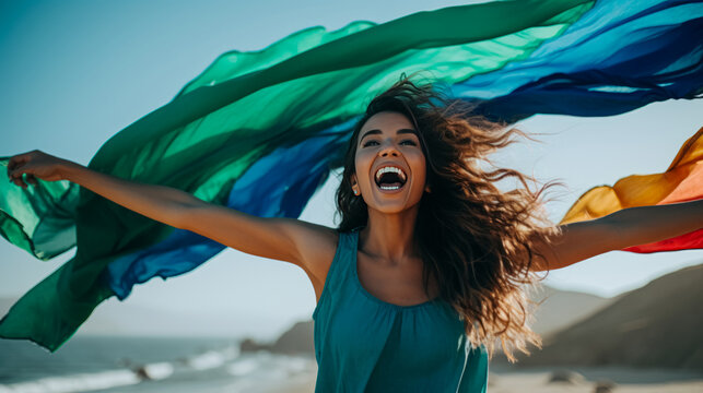 Joyful woman on the beach with arms outstretched, a colorful rainbow scarf flowing in the wind, with the ocean and blue sky in the background.