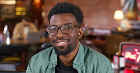 Portrait of African American man with glasses for better vision smiling positively while looking straight at camera. Young guy having recreation in modern cafe. In blurred background visible clients.