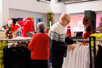 Senior people searching for jackets in shopping mall, looking at clothing items on hangers during...