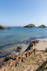 A tranquil beach scene with clear skies, rocky shore, calm waters, and distant green islands under a bright sky