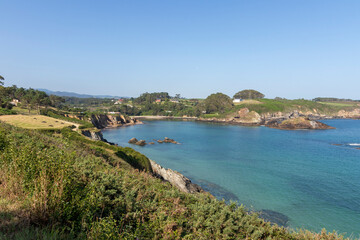 A scenic coastal view with clear blue waters, greenery, distant houses, and a bright, cloudless sky