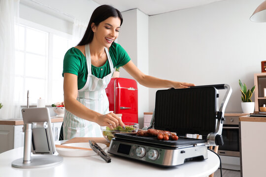 Young woman cooking sausages on electric grill in kitchen