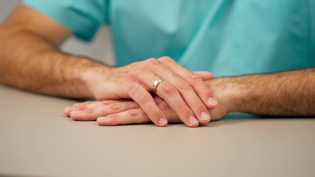 Close-up shot of a doctor's hands with a ring on his finger clasped together at a table and listening to patient