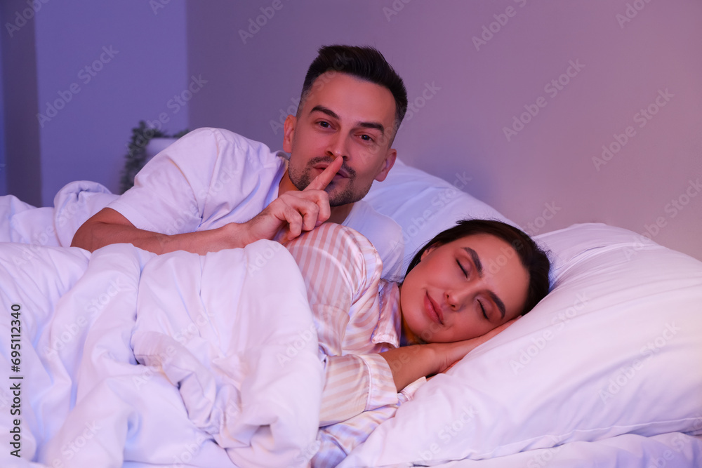 Wall mural Young man with his sleeping wife showing silence gesture in bedroom at night