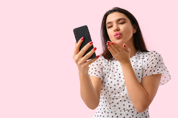 Young woman with mobile phone blowing kiss on pink background. Online dating concept
