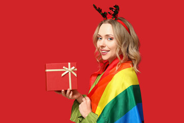 Beautiful young woman in reindeer horns with LGBT flag and gift box on red background. Christmas...