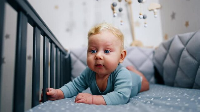 Blond Caucasian infant boy in blue bodysuit lies on the belly in the crib. Adorable blue-eyed boy looking up with surprise.