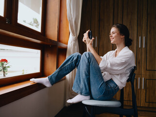 Young woman sitting by the window with phone in hand smile looking online, spring mood, home simple clothes, home cozy atmosphere, aesthetic lifestyle.