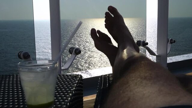 Relaxing on a cruise ship balcony with a refreshing cocktail overlooking the calm ocean sunset. A person rests their feet on a balcony railing while enjoying a cold drink during a peaceful vacation