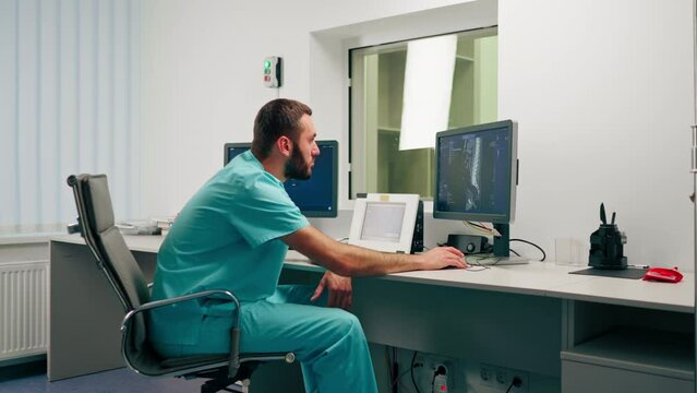 Concentrated male physician sits at a computer monitor in a room for describing MRI images and monitoring patient's condition