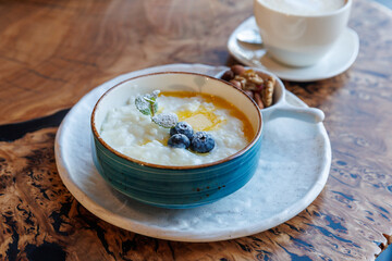 Milk rice porridge with blueberry, butter, nuts and mint leaf, creamy rice pudding or french riz au lait in a ceramic bowl with cup of tea or coffee on wooden table. Top view