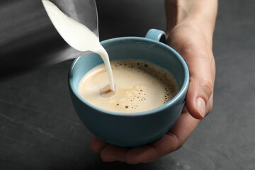 Woman pouring milk into cup of coffee at black table, closeup