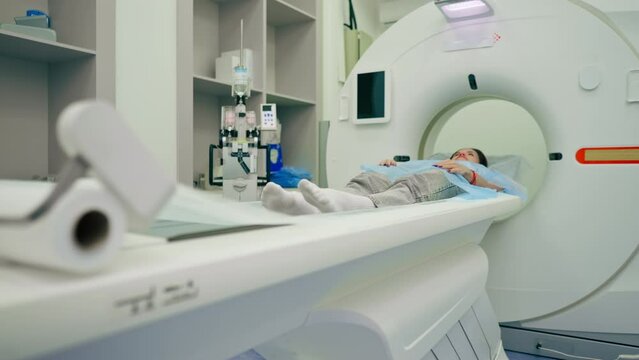 A girl in a medical gown lies on a movable couch of a magnetic resonance imaging machine and prepares for diagnosis