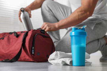 Young man putting bottle of water into bag indoors, focus on protein shake and towel
