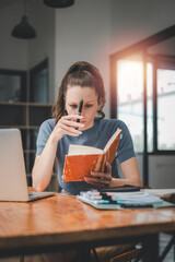 Obraz premium Business woman sitting at office desk and writing notes in a notebook while talking on a video call meeting with clients or coworkers on her laptop computer.