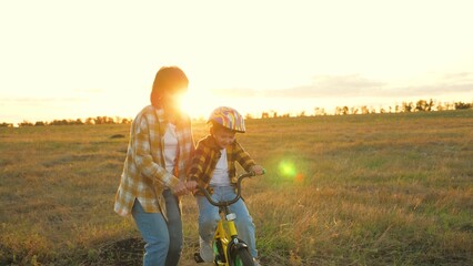 Optimistic boy practices cycling with support of patient mother in sunny evening. Little child and mommy relish beauty of nature engaging in biking. Young mom coaches cheerful son to ride bike