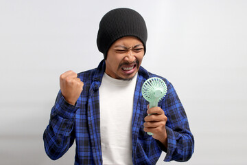 Angry Asian man, wearing a beanie hat and casual shirt, shouts and yells while holding a handheld portable mini fan, frustrated by the intense heat of the summer weather, isolated on white background © Jamaludinyusup