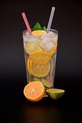 A tall glass of refreshing lemonade with ice and citrus pieces on a black background, next to ripe lime and orange slices.