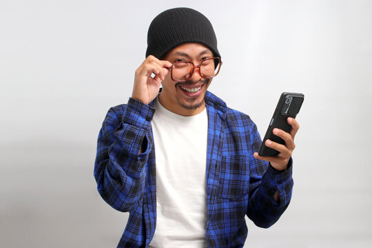 Asian man, dressed in a beanie hat and casual shirt, adjusts his eyeglasses and peeks above them, smiling as he looks at the camera with a mobile phone in hand, while standing against white background