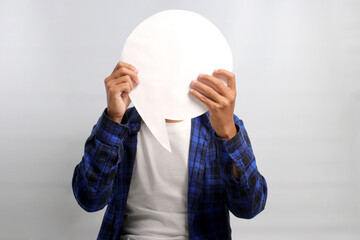 Asian man, concealing his face behind a speech bubble held in front of him, stands against a white background, depicting the concepts of information, thoughts, quotes, opinions, reviews, and feedback