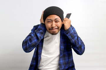 A discontented Asian man, dressed in a beanie hat and casual shirt, covers his ears in response to loud noises while holding a TV remote, isolated on a white background