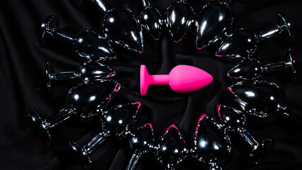 Pink silicone butt plug among many metal ones on a black silk sheet. 
