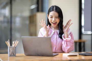 Photo of cheerful joyful young asian business woman in the office working on laptop digital project sitting at desk.
