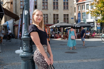 The photograph features a stylish young woman with blonde hair, standing confidently by a classic street lamp. She is dressed in a chic black top paired with a zebra print skirt, which adds a trendy