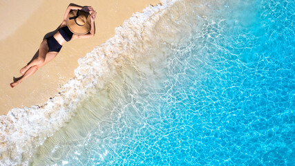 Aerial view of beautiful young woman in black swimsuit lying on sandy beach near blue sea with...