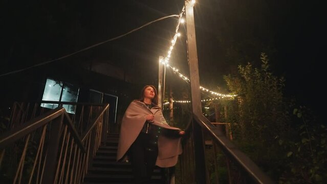 Contemplative lady descends stairs in backyard. Pensive woman strolls through well-lit private spaces of home late in evening. Bright illumination